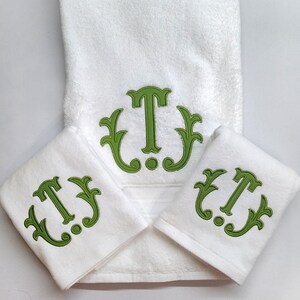 May include: A white towel set featuring a large green embroidered letter "T" with decorative flourishes. The set includes a large bath towel and two smaller hand towels, all with the same monogram design.