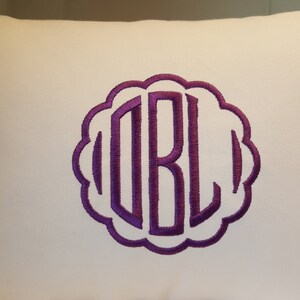 May include: White pillow with a purple embroidered monogram of the letters "DBL" in a decorative circle.