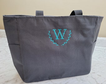 May include: A gray canvas tote bag with a teal monogram of the letter "W" in a wreath design.