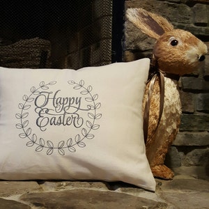 May include: A white pillow with the text "Happy Easter" embroidered in a wreath of leaves. The pillow is sitting on a stone surface next to a brown bunny figurine.