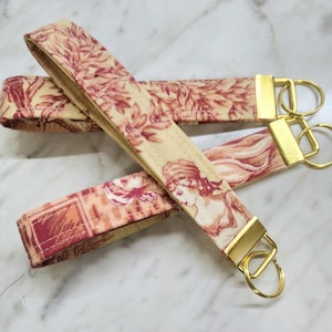 May include: Two fabric keychains with gold key rings. The keychains are cream colored with a red floral pattern. One keychain has a gold ring with two loops, the other has a gold ring with one loop.