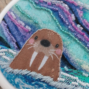 May include: A brown and white embroidered walrus with a pink nose and black eyes, surrounded by blue and purple fabric with white beads. The fabric is stitched into a circular frame.