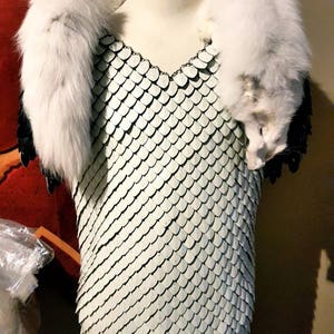 May include: A white chainmail shirt with a white fur collar. The fur collar is draped over the shoulder of a mannequin.