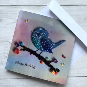 May include: A handmade birthday card featuring a blue beaded bird perched on a branch with brown buttons and colorful flowers. The card has a watercolor background and the text "Happy Birthday" is written in black.
