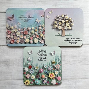 May include: Three square coasters with a floral design and inspirational quotes. The first coaster features a blue and pink background with a field of flowers and the quote "For I know the plans I have for you, plans to give you hope and a future. Jr 29:11". The second coaster features a light blue background with a tree made of buttons and the quote "Don't worry about anything, but pray about everything. Ph 4:6-7". The third coaster features a light green background with a field of flowers and the quote "He restores my soul. Psalm 23:3".