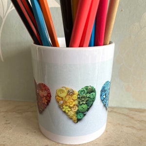 May include: White ceramic mug with a rainbow heart design made of colorful buttons. The heart is made of red, yellow, and green buttons. The mug is filled with colorful pencils.