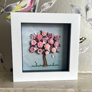 May include: A framed artwork depicting a tree with a brown trunk and a full crown of pink flowers. The flowers are in various shades of pink, creating a blossoming effect. The artwork is set against a light blue background and framed in a white square frame.
