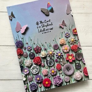 May include: A decorative canvas art piece with a pastel sky background, butterflies, and floral embellishments. The artwork includes the text "The Lord is my Shepherd I shall not want" and is decorated with buttons and other elements.