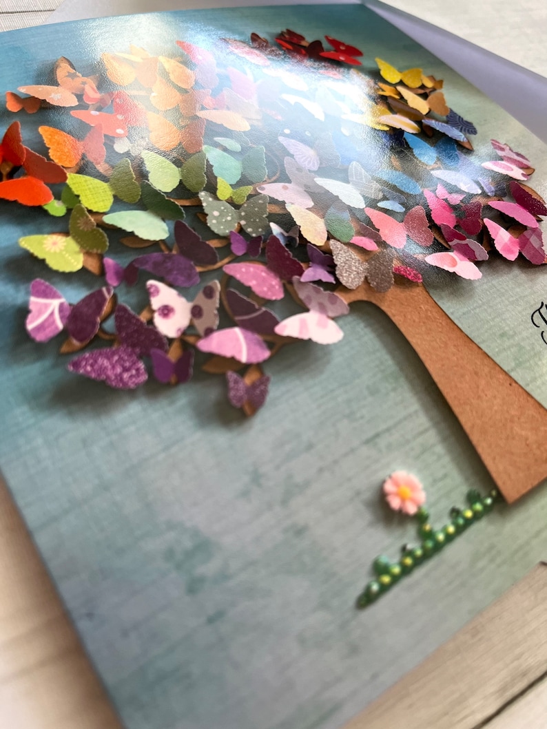 May include: A colorful card with a 3D paper butterfly tree. The tree is made of many butterflies in shades of purple, blue, green, yellow, orange, and red. A small pink flower with green leaves is at the base of the tree.