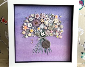 May include: A framed artwork featuring a bouquet made of buttons and beads in shades of pink, white, and cream, set against a purple background. The bouquet includes a silver charm with the text "Sometimes you forget that you're awesome".