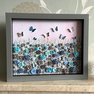 May include: A framed artwork featuring a blue and grey button flower field with butterflies flying above. The background is a light pink colour.