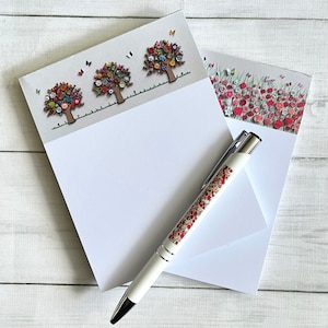 May include: Two white memo pads with a floral design. The top pad features three colourful trees with buttons for leaves. The bottom pad has a field of pink and red flowers. A white pen with a floral design is on top of the pads.