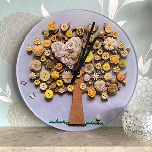 May include: A purple clock with a tree design made of buttons. The tree has yellow, white, and brown buttons. The clock hands are black.