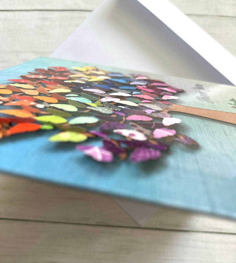 May include: A greeting card with a blue background and a colorful butterfly tree design. The tree is made of paper butterflies in various colors and patterns. The card has the text "Happy Day" on the front.