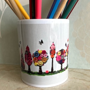 Peut inclure: Une tasse en céramique blanche avec un motif d'arbre à boutons colorés. Les arbres sont faits de boutons de différentes couleurs et ont des troncs noirs. De petits papillons noirs volent autour des arbres. La tasse est remplie de crayons.
