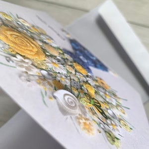 May include: A handmade greeting card with a 3D floral design. The flowers are made of white, yellow, and blue buttons and are arranged in a bouquet. There is a small green butterfly and a seashell in the bouquet.