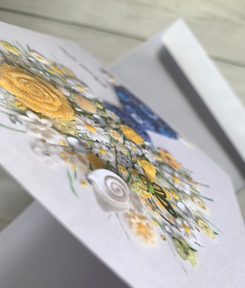 May include: A handmade greeting card with a 3D floral design. The flowers are made of white, yellow, and blue buttons and are arranged in a bouquet. There is a small green butterfly and a seashell in the bouquet.