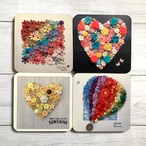 May include: Four square coasters with different designs made of buttons. The first coaster has a heart shape made of pink, red, and blue buttons with the text "be courage and kind". The second coaster has a heart shape made of pink, red, and yellow buttons with the text "follow your dreams". The third coaster has a heart shape made of yellow and white buttons with the text "always bring your own sunshine". The fourth coaster has a hot air balloon shape made of rainbow colored buttons.
