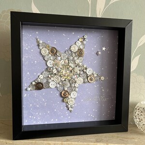 May include: A framed artwork featuring a large star made of buttons in shades of white, silver, and gold on a light purple background with white stars. The text "You are my shining star" is written below the star.