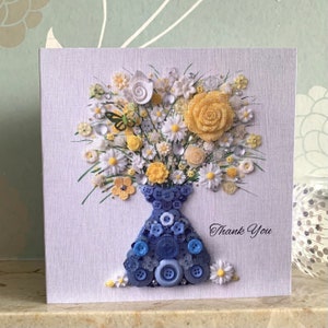 May include: A handmade card with a blue button vase filled with white and yellow flowers. The card has the words "Thank You" written on it.