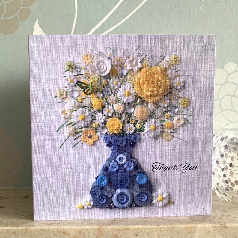 May include: A handmade card with a blue button vase filled with white and yellow flowers. The card has the words "Thank You" written on it.