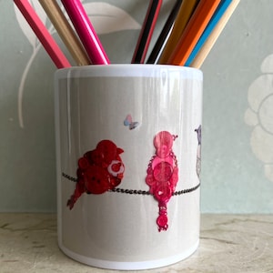 May include: A white ceramic pencil holder featuring a design of three birds made from buttons on a wire. The birds are red, pink, and grey. There is a small butterfly in the background.