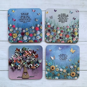 May include: Set of four square coasters with inspirational bible verses printed on them. Each coaster features a different design with a blue background and colorful flowers, butterflies, and buttons. The verses are: "The Lord is my light and my salvation; whom shall I fear? The Lord is the strength of my life; of whom shall I be afraid?" (Psalm 27:1), "The Lord will guide you continually and satisfy your soul in drought, and you will be like a watered garden, and like a spring of water whose waters fail not." (Isaiah 58:11), "The Lord will fight for you; you need only to be still." (Exodus 14:14), and "My grace is sufficient for you, for my power is made perfect in weakness." (2 Corinthians 12:9).