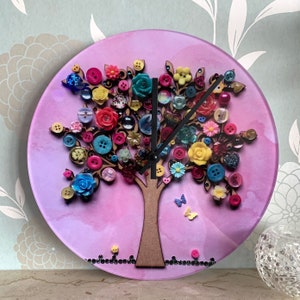 May include: A pink clock with a tree design made of buttons. The tree has colourful buttons, flowers, and leaves. The clock hands are black.