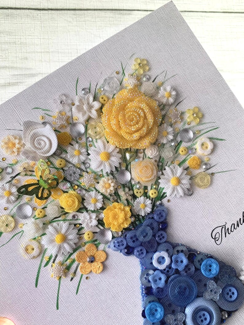 May include: A handmade card featuring a bouquet of yellow and white flowers in a blue button vase. The flowers are made of fabric and buttons, and the vase is made of buttons. The card has the word "Thank" written on it.