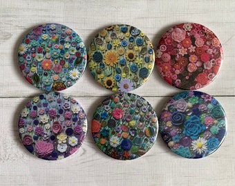May include: Six round decorative magnets with floral designs. Each magnet features a unique arrangement of flowers and buttons in various colors, including blue, pink, yellow, and purple. The magnets are arranged on a white wooden surface.