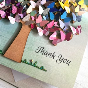 May include: A handmade card with a paper tree decorated with colorful butterflies. The tree is brown and the butterflies are in shades of pink, purple, blue, green, yellow, and orange. The card has a green background and the words "Thank You" are written in black cursive.