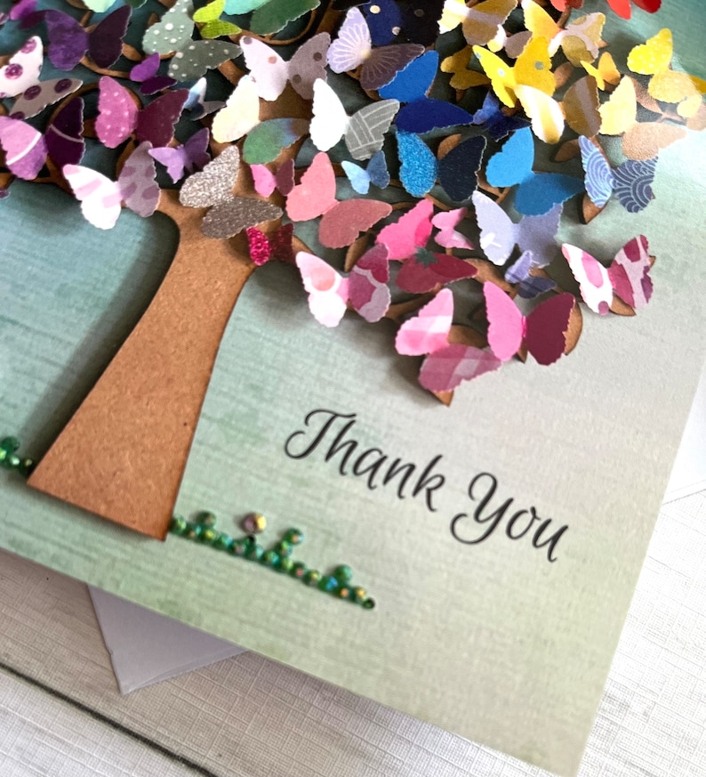 May include: A handmade card with a paper tree decorated with colorful butterflies. The tree is brown and the butterflies are in shades of pink, purple, blue, green, yellow, and orange. The card has a green background and the words "Thank You" are written in black cursive.
