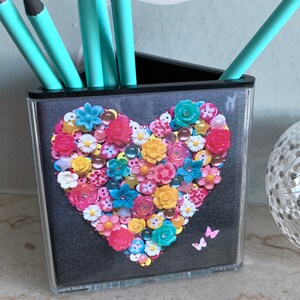 May include: A black triangular desk organiser with a heart-shaped design made of colourful buttons. The heart is surrounded by a black background and the organiser has three compartments for pens and pencils.
