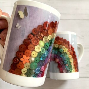 May include: Two white ceramic mugs featuring a rainbow design made from colourful buttons. The mugs have a handle on the right side. The background is a light wood surface.