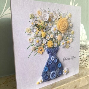 May include: A handmade card featuring a bouquet of yellow and white flowers made from buttons, arranged in a blue button vase. The card has the words "Thank You" written on it.