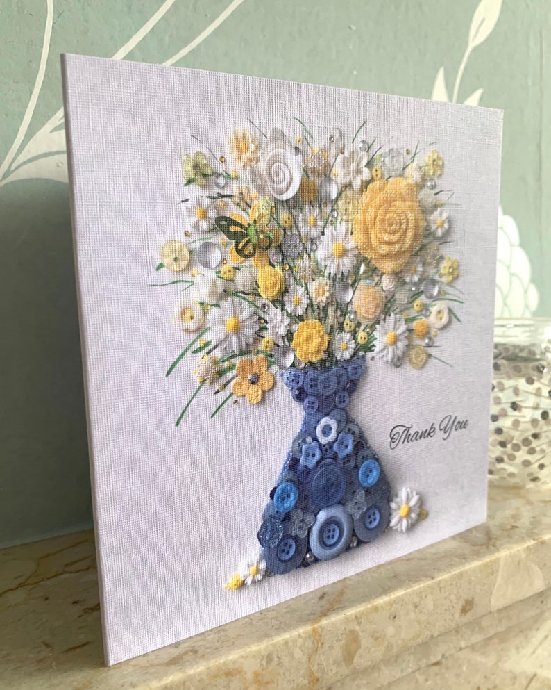 May include: A handmade card featuring a bouquet of yellow and white flowers made from buttons, arranged in a blue button vase. The card has the words "Thank You" written on it.