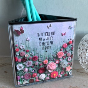 May include: A triangular clear acrylic pencil holder with a decorative design featuring a field of flowers, buttons, and butterflies. The design includes the text "To the world you are a mother, to me you are the world."