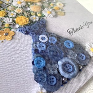 May include: A handmade thank you card featuring a bouquet of yellow and white flowers in a blue button vase. The card has a white background with the words "Thank You" written in black.