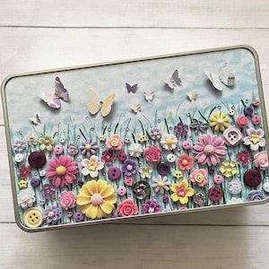 May include: A rectangular metal tin with a floral and butterfly design. The lid features a painted scene of colorful flowers, butterflies, and buttons. The flowers are in shades of pink, yellow, purple, and white, with green grass and a blue sky background.