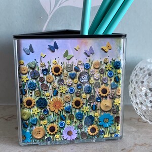 May include: A black triangular desk organiser with a colourful floral design. The design features a field of yellow sunflowers, blue flowers, and green leaves with a variety of colourful buttons scattered throughout. The design also includes butterflies in shades of blue and purple.