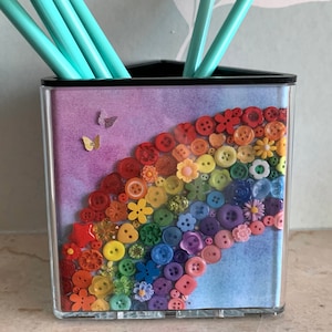 May include: A triangular clear pencil holder with a rainbow design made of colorful buttons and flowers. The background is a watercolor wash of purple, blue, and white.