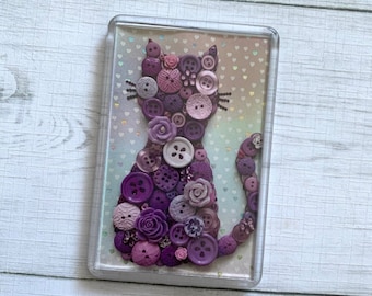 May include: A purple cat silhouette made of buttons, with a glittery background. The cat is made of various shades of purple buttons, including some with floral designs.