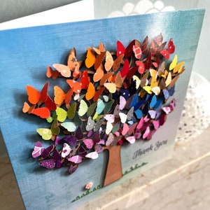 May include: A greeting card with a colorful butterfly tree design. The tree is made of paper butterflies in shades of red, orange, yellow, green, blue, purple, and pink. The card has the words "Thank You" written on it.