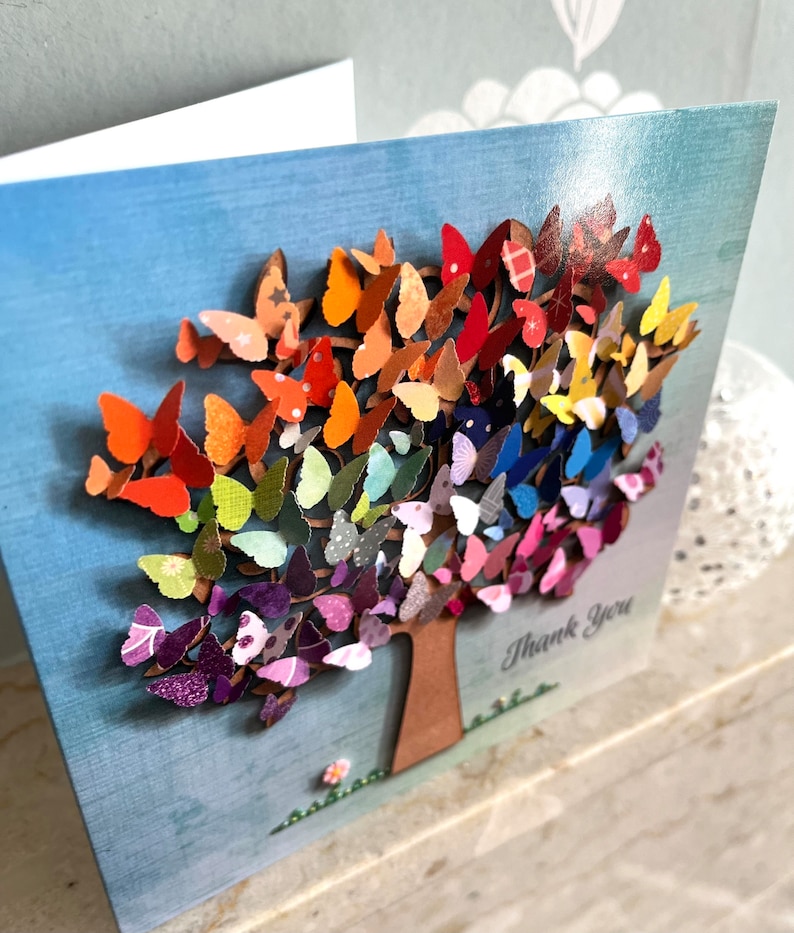 May include: A greeting card with a colorful butterfly tree design. The tree is made of paper butterflies in shades of red, orange, yellow, green, blue, purple, and pink. The card has the words "Thank You" written on it.