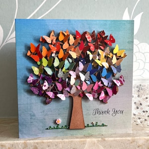 May include: A handmade card featuring a colorful paper butterfly tree with a brown wooden trunk and the words "Thank You" written in black script.