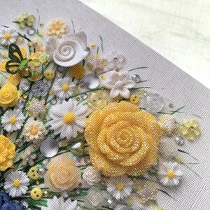 May include: A close-up of a yellow and white floral arrangement made with fabric flowers, buttons, and beads. The arrangement is on a gray canvas background.