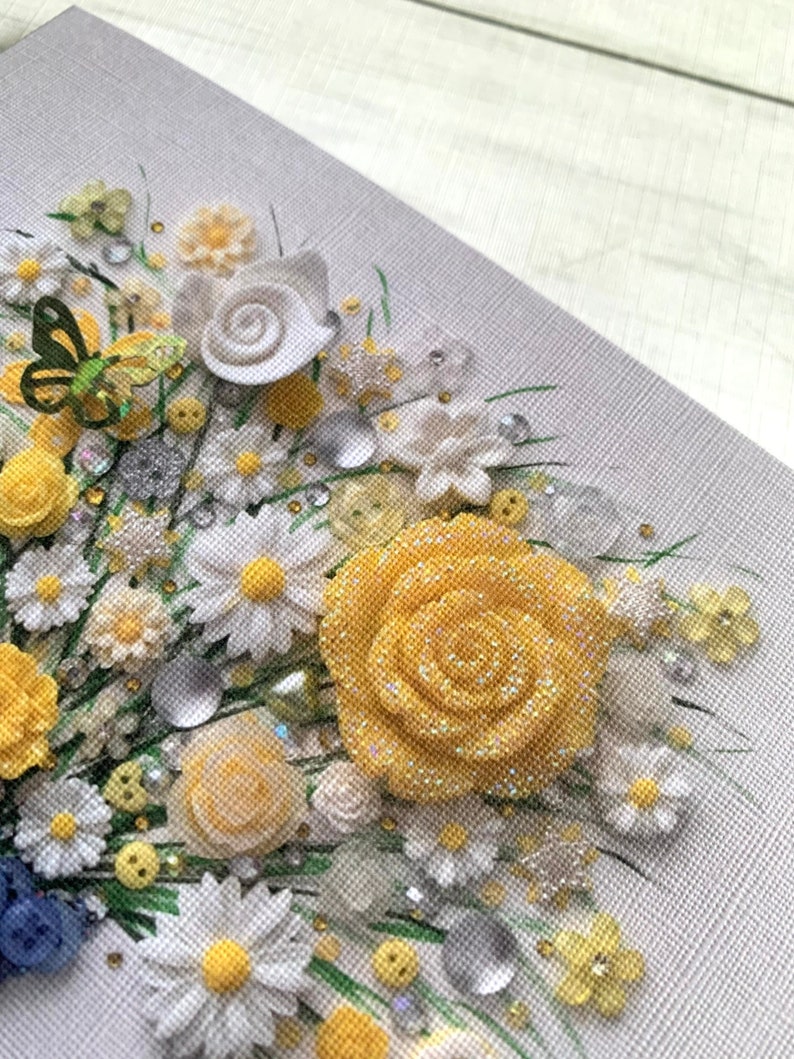 May include: A close-up of a yellow and white floral arrangement made with fabric flowers, buttons, and beads. The arrangement is on a gray canvas background.