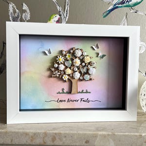 May include: A framed picture with a tree design, decorated with flowers and butterflies, set against a pastel watercolour backdrop. The tree has white, yellow, and cream flowers. The words "Love Never Fails" are written below.