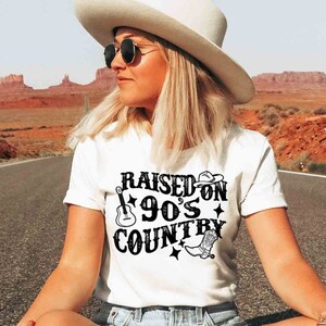 K&ouml;nnte beinhalten: Wei&szlig;es T-Shirt mit schwarzem Text, der "Raised on 90's Country" lautet. Der Text ist in einer Retro-Schriftart mit einem Gitarren- und Cowboystiefel-Motiv. Das Shirt wird von einer Person getragen, die auf einer Stra&szlig;e mit einer W&uuml;stenlandschaft im Hintergrund sitzt.