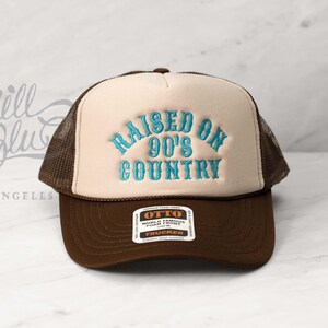 May include: A brown and tan trucker hat with the text "RAISED ON 90'S COUNTRY" embroidered in turquoise thread. The hat features a mesh back and a curved brim. The brand tag "OTTO" is visible on the front.